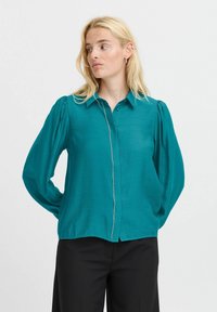 Camicia azzurro-turchese con taglio dritto, maniche a sbuffo e una finitura decorativa in argento lungo il fronte. Tessuto morbido e leggero.