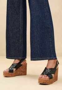 Sandali neri intrecciati con tacco in sughero, caratterizzati da un design con fasce incrociate e una fibbia alla caviglia. Abbinati a jeans a gamba larga in denim scuro.