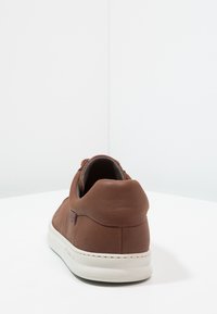 Zapatilla de cuero marrón con una textura suave, parte trasera redondeada, detalles de costura sutiles y una suela de goma blanca.