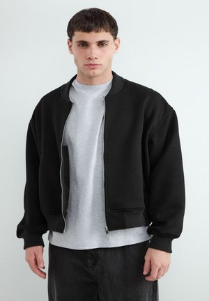 Jeune homme portant une veste bomber noire sur un t-shirt gris clair et un pantalon foncé, debout devant un fond clair uni.