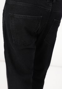 Jeansi din denim negri cu un design ajustat, având două buzunare pe spate și detalii subtile de cusătură, surprinși dintr-un unghi lateral.
