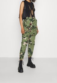 Sort ærmeløs top med udskæringer, parret med grønne camouflage cargobukser med en bølget mønster og flere lommer. Sorte klodsede støvler.