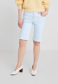 Bermudas de mezclilla azul claro con un cierre de cinco botones, bolsillos frontales y un corte relajado. Combinadas con un suéter de punto amarillo y tacones blancos.