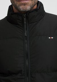 Črna podložena jakna z visokim ovratnikom, celotnim zadrgo in majhnim tridelnim logotipom na prsih. Mat tekstura in šivana obdelava.