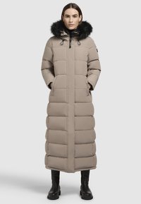 Hosszú bézs színű puffermellény, lecipzározható fekete szőrme kapucnival, megkötős derékkal, oldalsó zsebekkel és egyenes fazonnal. Fekete csizmák láthatók.