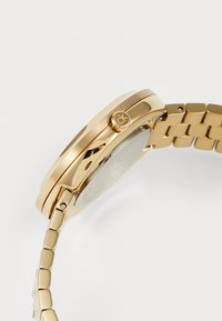 Gold wristwatch with a smooth metal case, textured band links, and a side button featuring a logo. Reflective polished surface detail.