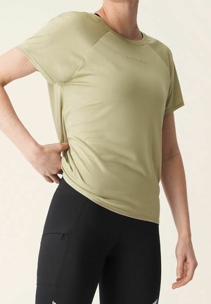 Person trägt ein hellgrünes kurzärmeliges Shirt und schwarze Sportleggings, Hand auf der Hüfte, vor schlichtem Hintergrund.