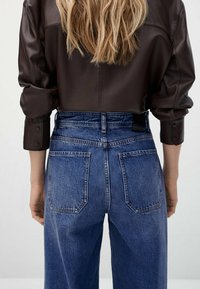 Chemise en cuir marron, jean bleu en denim taille haute avec quatre poches, légère décoloration, coutures visibles, tissu texturé, coupe décontractée.