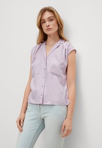 Lavender short-sleeve blouse with a collar, two chest pockets, and button closure. Smooth fabric with a relaxed fit and side slits.