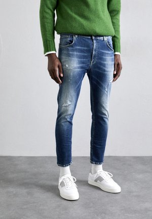 Personne portant un pull vert, un jean bleu déchiré, des chaussettes blanches et des baskets blanches, debout sur un sol gris contre un mur uni.