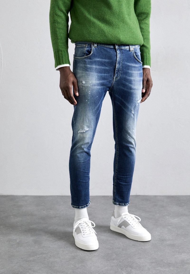 Personne portant un pull vert, un jean bleu déchiré, des chaussettes blanches et des baskets blanches, debout sur un sol gris contre un mur uni.