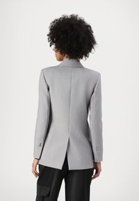 Grå blazer med en strukturert silhuett, klaffelapp og enkel vent bak. Har metalliske detaljer på ermene og glatt tekstur.