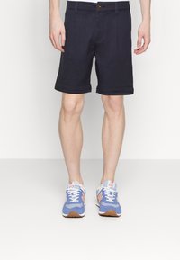 Man som bär mörkblå shorts och blå, persiko- och vita träningsskor, stående mot en vit bakgrund.