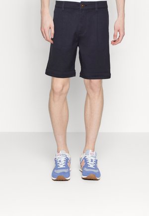 Homme portant un short bleu marine et des baskets de sport bleu, pêche et blanc, se tenant devant un fond blanc.