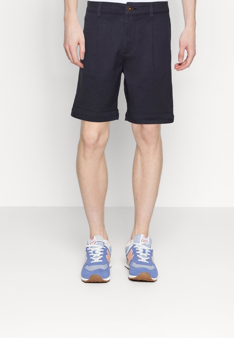 Man som bär mörkblå shorts och blå, persiko- och vita träningsskor, stående mot en vit bakgrund.