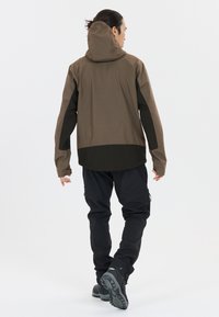Braune wasserdichte Jacke mit passgenauer Kapuze, schwarzen Akzenten, langen Ärmeln und lockerer Passform, kombiniert mit schwarzen Hosen und strukturiertem Schuhwerk.
