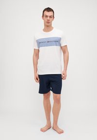 Bijela majica s plavim horizontalnim prugama koja sadrži logo "TOMMY HILFIGER"; uz nju su kombinirane tamnoplave šortseve od glatkog materijala, opuštenog kroja.