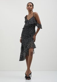 Schwarzes Kleid mit Punktemuster, Rüscheneinsatz, asymmetrischem Saum, Schulterdesign mit kaltem Look, leichtem Stoff und taillierter Silhouette.