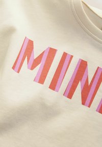 Tissu beige avec de grandes lettres rayées en rouge et rose formant une partie du mot "MINN" sur un vêtement en gros plan.