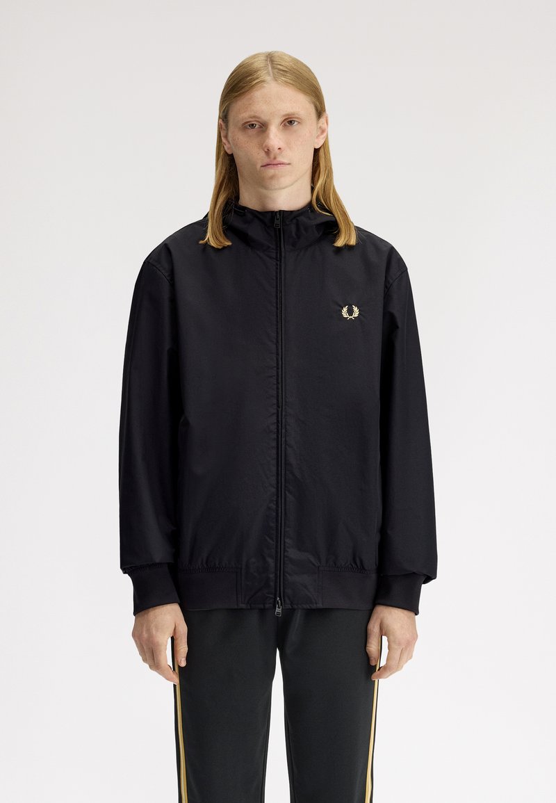 Persona joven con cabello largo y liso que lleva una chaqueta negra con cremallera y pantalones negros con rayas doradas, de pie contra un fondo claro y sencillo.