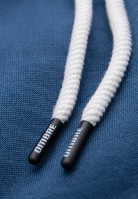 Close-up af hvide flettede snore til hættetrøje med sorte plastikender mærket "Dimbre" på en blå stofbaggrund.