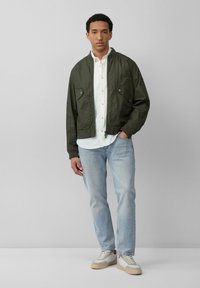 Veste bombardier olive avec deux poches à pression, portée par-dessus une chemise blanche à boutons. Un jean en denim bleu clair et des baskets beiges complètent le look.