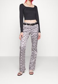 Kvinna som bär en svart långärmad crop top, byxor med ormskinnsmönster och ett svart bälte, guldhalsband och svarta klacksandaler.