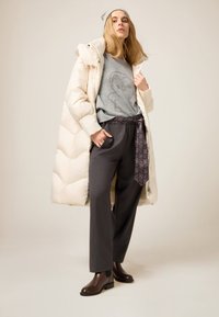 Cappotto crema imbottito sopra maglione grigio con motivo decorativo, pantaloni grigi a gamba larga e stivaletti marroni; presenta tessuto decorato annodato in vita.
