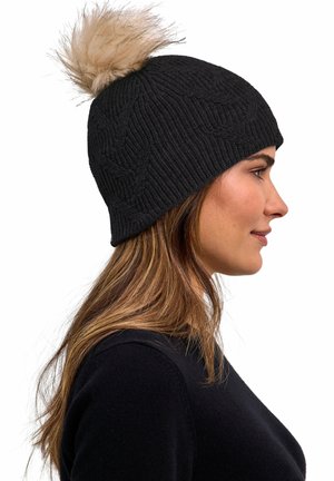 Frau mit schwarzer Strickmütze mit beigem Kunstpelzbommel, trägt schwarzen Pullover, im Profil vor weißem Hintergrund gezeigt.