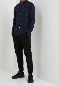 Camicia a maniche lunghe a quadri blu navy con collo a fascia, con bottoni bianchi; abbinata a pantaloni neri a gamba stretta e sneaker nere.