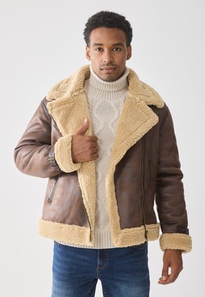 Chaqueta de cuero marrón con un cuello y puños de forro polar beige, que cuenta con un cierre de cremallera y un cinturón decorativo en la manga. Llevada sobre un suéter de punto.