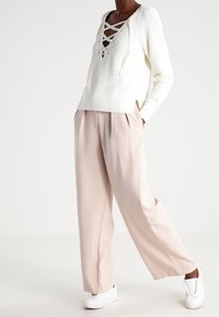 Witte gebreide trui met V-hals en veterdetail, gecombineerd met lichtroze, relaxte broek en witte sneakers.