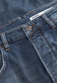 Pantalones vaqueros de mezclilla azul con costuras naranjas, un botón metálico y una etiqueta con la palabra "GABBA". Presentan un diseño tradicional de cinco bolsillos.