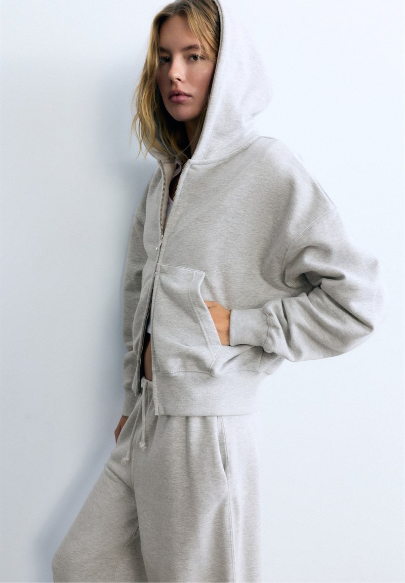 Femme portant un sweat à capuche zippé gris clair et un pantalon de jogging assorti, une main dans la poche, sur un fond uni.