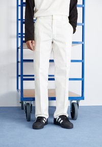 White trousers with a straight cut, featuring a smooth texture. Paired with black sneakers with white stripes. Background includes blue shelving.