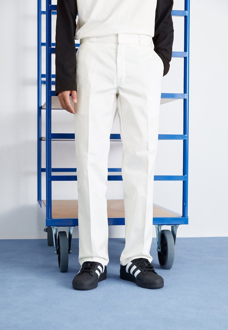 White trousers with a straight cut, featuring a smooth texture. Paired with black sneakers with white stripes. Background includes blue shelving.