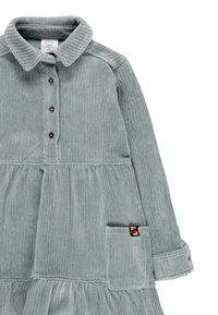 Vestito in corduroy azzurro chiaro con colletto, chiusura frontale a bottoni, maniche lunghe e orlo arricciato. Include una tasca a toppa con un disegno floreale.