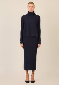 Navy Pullover mit hohem Rollkragen, gerippter Textur und Knopfdetails an den Ärmeln, kombiniert mit einem passenden gerippten Midirock. Hohe Absätze.