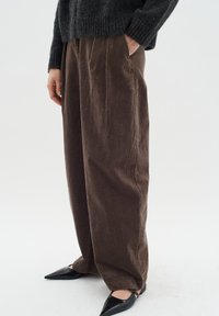 Pantaloni in velluto a coste marroni con un design a gamba larga, dotati di pieghe frontali e tasche laterali. Abbinati a scarpe nere a punta.