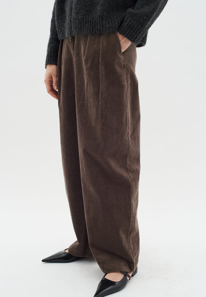 Pantaloni in velluto a coste marroni con un design a gamba larga, dotati di pieghe frontali e tasche laterali. Abbinati a scarpe nere a punta.