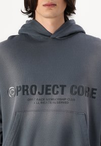 Grauer Kapuzenpullover mit einer Vordertasche. Text "PROJECT CORE" und "OFF-TRACK MEMBERSHIP CLUB" in dunklen, fetten Buchstaben aufgedruckt. Weicher Stoff.