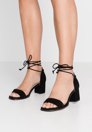 Czarne zamszowe sandały na obcasie z wiązaniami wokół kostki, otwarty przód, słupkowy obcas i podwójne paski z przodu. Gładka faktura i minimalne ozdobniki.