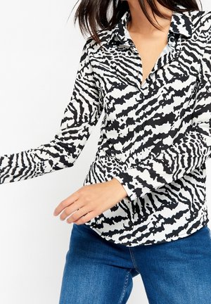 Kvinde iført en sort-hvid zebra-stribet skjorte med lange ærmer og blå jeans med den ene arm strakt ud til siden.