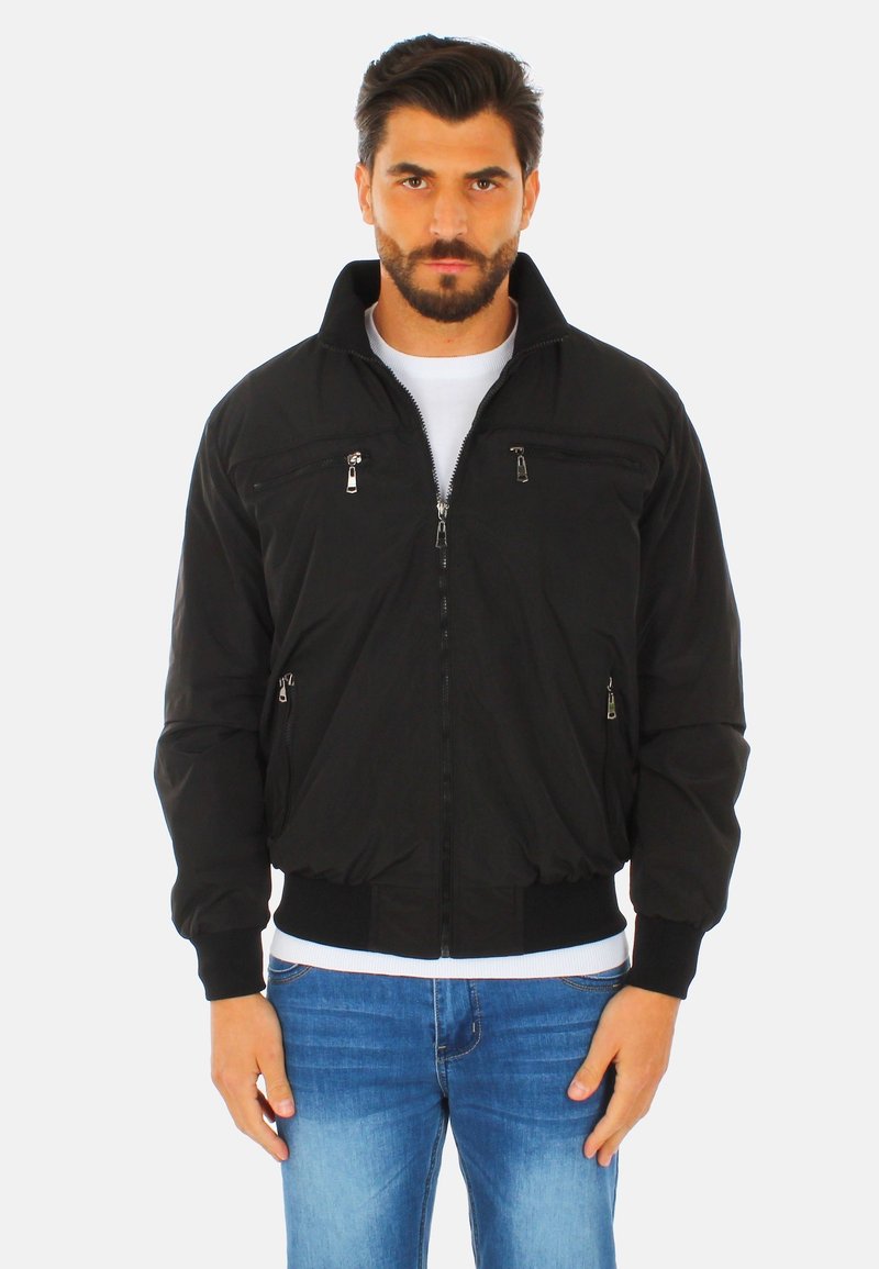 Giubbotto bomber nero con tasche a zip, colletto alto e polsini a coste. Il tessuto è liscio; il giubbotto presenta un design aderente. Indossato sopra una camicia bianca.