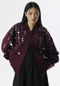 Chaqueta bomber burdeos con una textura suave, adornada con lentejuelas plateadas en patrones verticales y puños acanalados, combinada con un vestido negro.