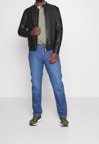 Black leather bomber jacket with no collar, green polo shirt, blue jeans, and green camo sneakers, standing against a plain background.