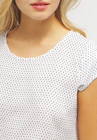 Bílá blůza s kulatým výstřihem, krátkými rukávy a jemným vzorem malých černých teček. Lehce látkové s hladkým povrchem.