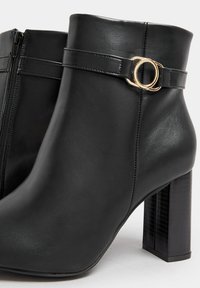 Botas de tobillo negras hechas de cuero suave, con un cierre lateral, tacón cuadrado y un acento decorativo en forma de bucle de metal dorado en la correa.