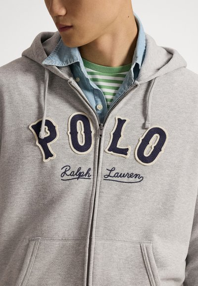 Άνδρας που φοράει γκρι φούτερ με φερμουάρ και το γράμμα "POLO Ralph Lauren" πάνω από πουκάμισο με πράσινες ρίγες και μπλε τζιν πουκάμισο με κουμπιά στο γιακά.