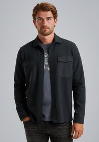 Temno siva srajca z gumbi in teksturiranim materialom, z dvema žepoma na prsih in ovratnikom, oblečena preko sivo majice in v kombinaciji z temno modrimi kavbojkami.